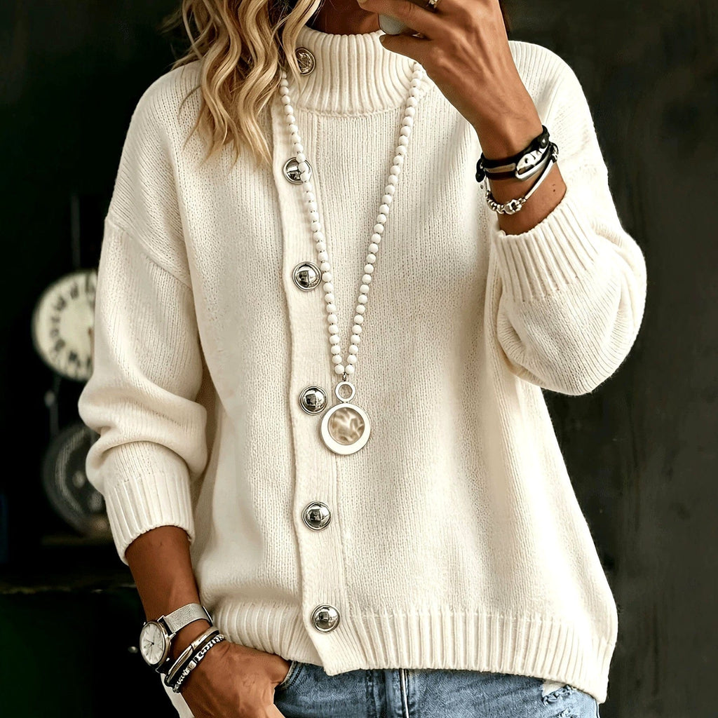 Woman wearing a cream mock neck knit sweater with silver domed buttons down the front, ribbed cuffs and hem, styled with blue jeans and a long beaded necklace._0