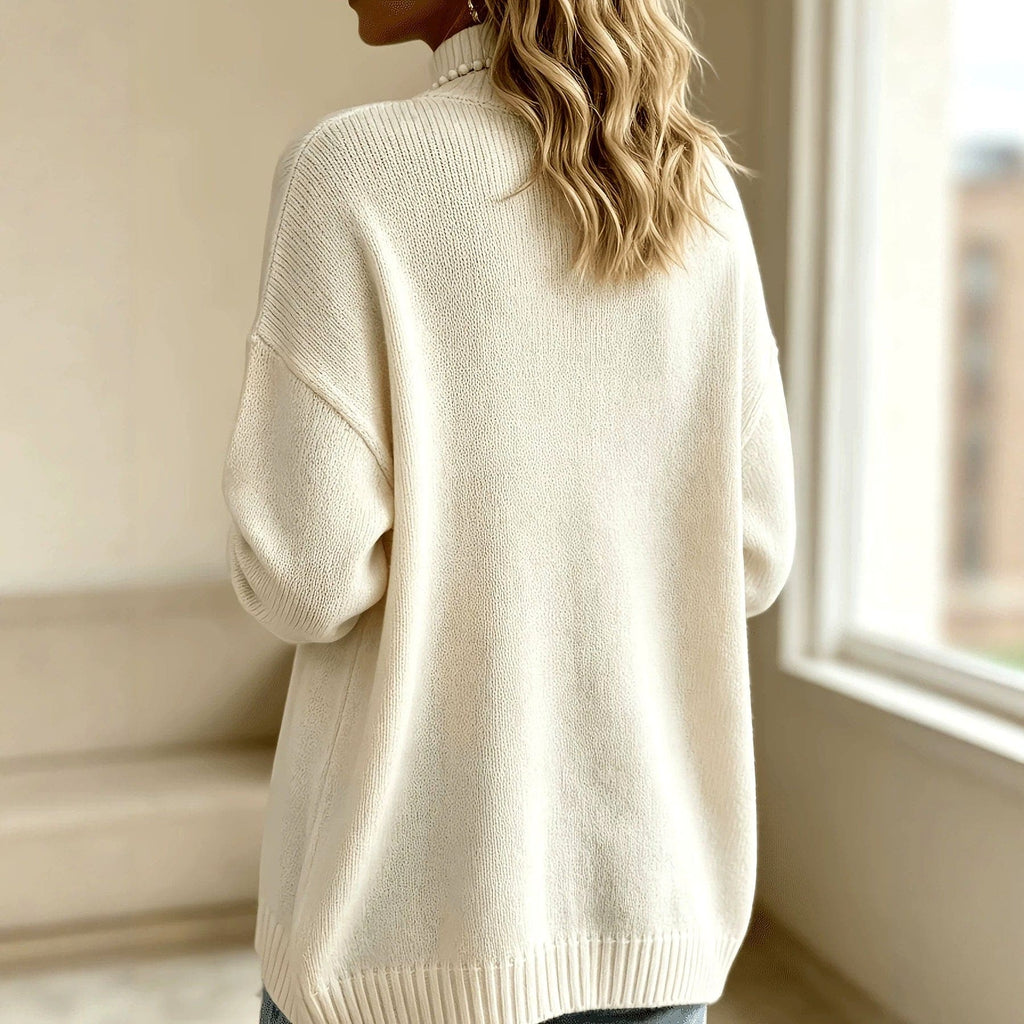Woman wearing a cream mock neck knit sweater with silver domed buttons down the front, ribbed cuffs and hem, styled with blue jeans and a long beaded necklace._2