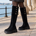 Pair of black knee-high boots with gold buckle straps, stud details and thick lugged platform soles, worn on a stone promenade by the water._0
