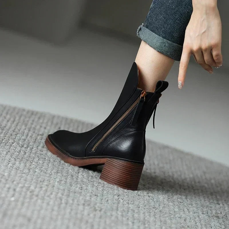 Black ankle boot worn with cuffed jeans, showing the side zipper, pointed toe, pull tab, and brown block heel from the side.