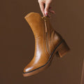 A hand holding a tan ankle boot by the zipper pull, showcasing the side zipper closure and block heel design._0