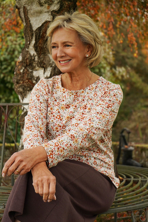 Smiling woman seated at a wooden table wearing a cream long sleeve blouse with a small orange and amber floral print and a round neckline._1