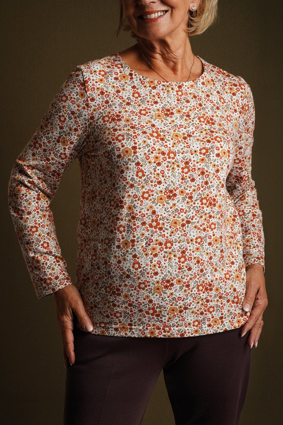 Smiling woman seated at a wooden table wearing a cream long sleeve blouse with a small orange and amber floral print and a round neckline._2