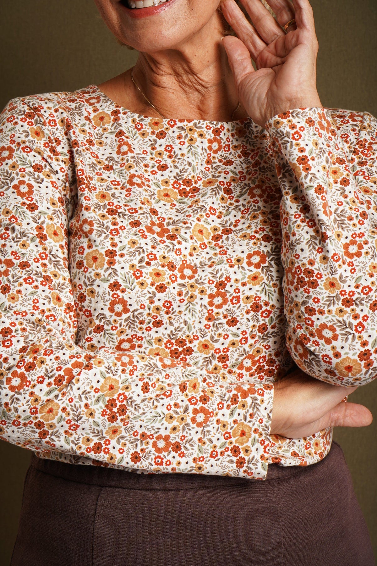 Smiling woman seated at a wooden table wearing a cream long sleeve blouse with a small orange and amber floral print and a round neckline._3