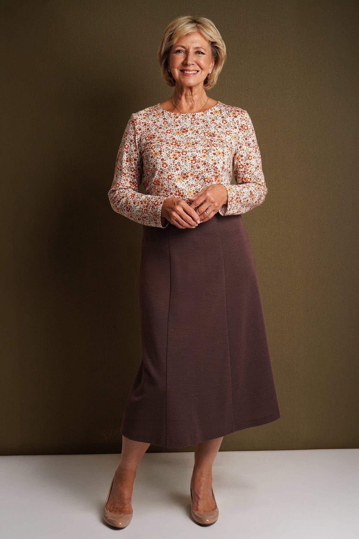 Smiling woman seated at a wooden table wearing a cream long sleeve blouse with a small orange and amber floral print and a round neckline._4