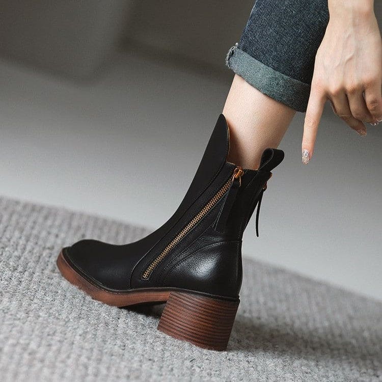 Pair of black mid-calf boots with gold side zips, brown stacked block heels and rounded toes on a white plinth._2
