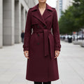 Woman wearing a belted double-breasted trench coat in deep burgundy with notched lapels and a tied waist belt, standing outdoors on a city sidewalk._0