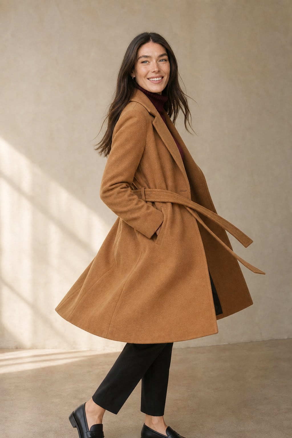 Woman wearing a camel belted midi coat with tie waist over a turtleneck and black trousers, standing in natural light._0