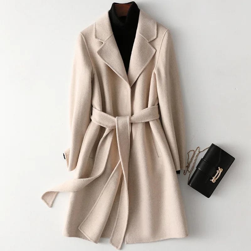 Woman wearing a camel belted midi coat with tie waist over a turtleneck and black trousers, standing in natural light._18