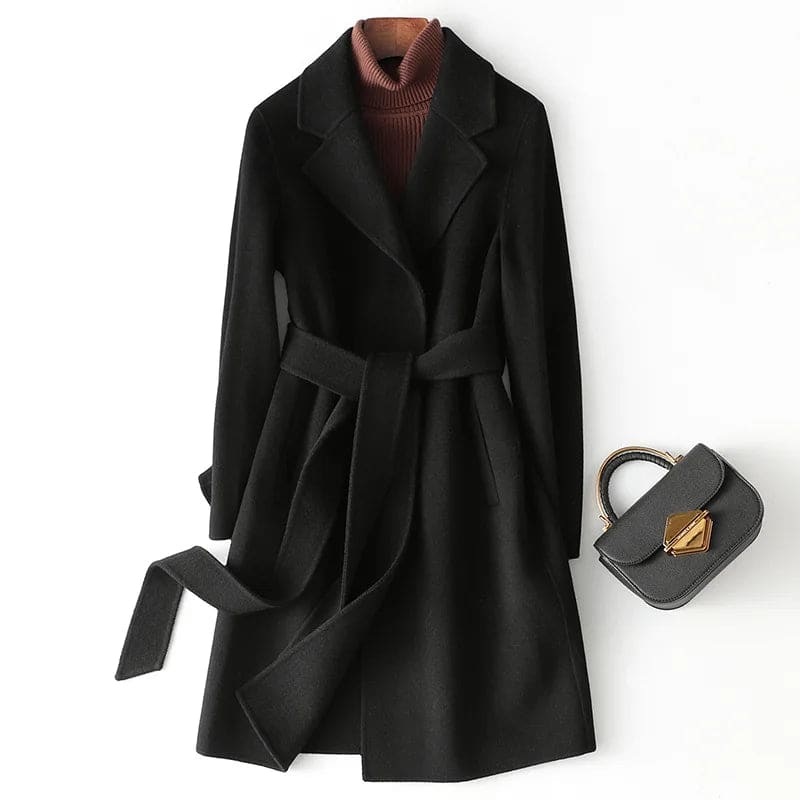 Woman wearing a camel belted midi coat with tie waist over a turtleneck and black trousers, standing in natural light._22