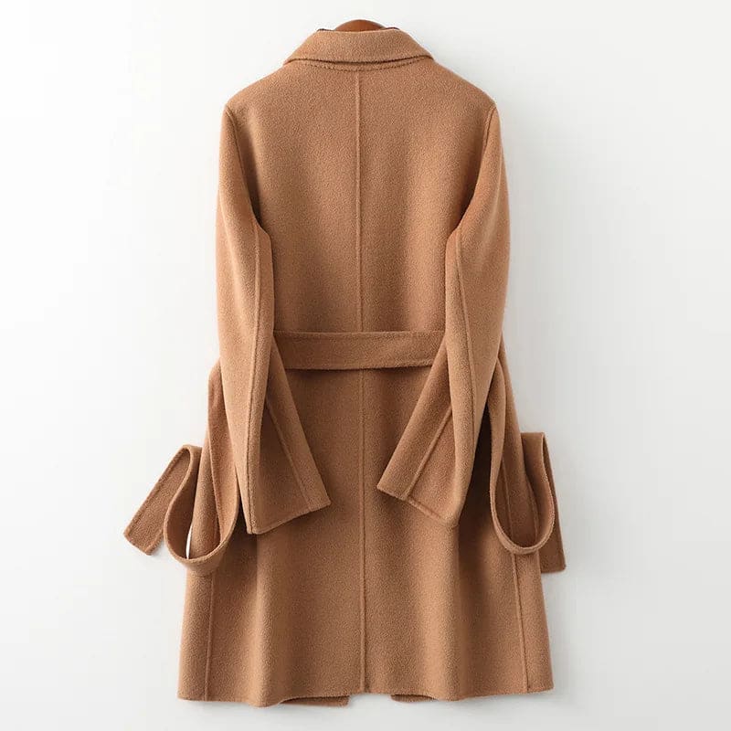 Woman wearing a camel belted midi coat with tie waist over a turtleneck and black trousers, standing in natural light._23