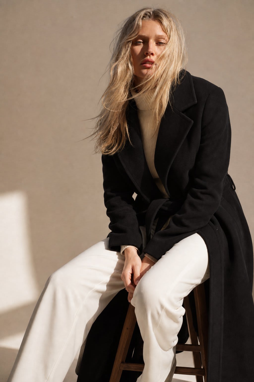 Woman wearing a camel belted midi coat with tie waist over a turtleneck and black trousers, standing in natural light._4