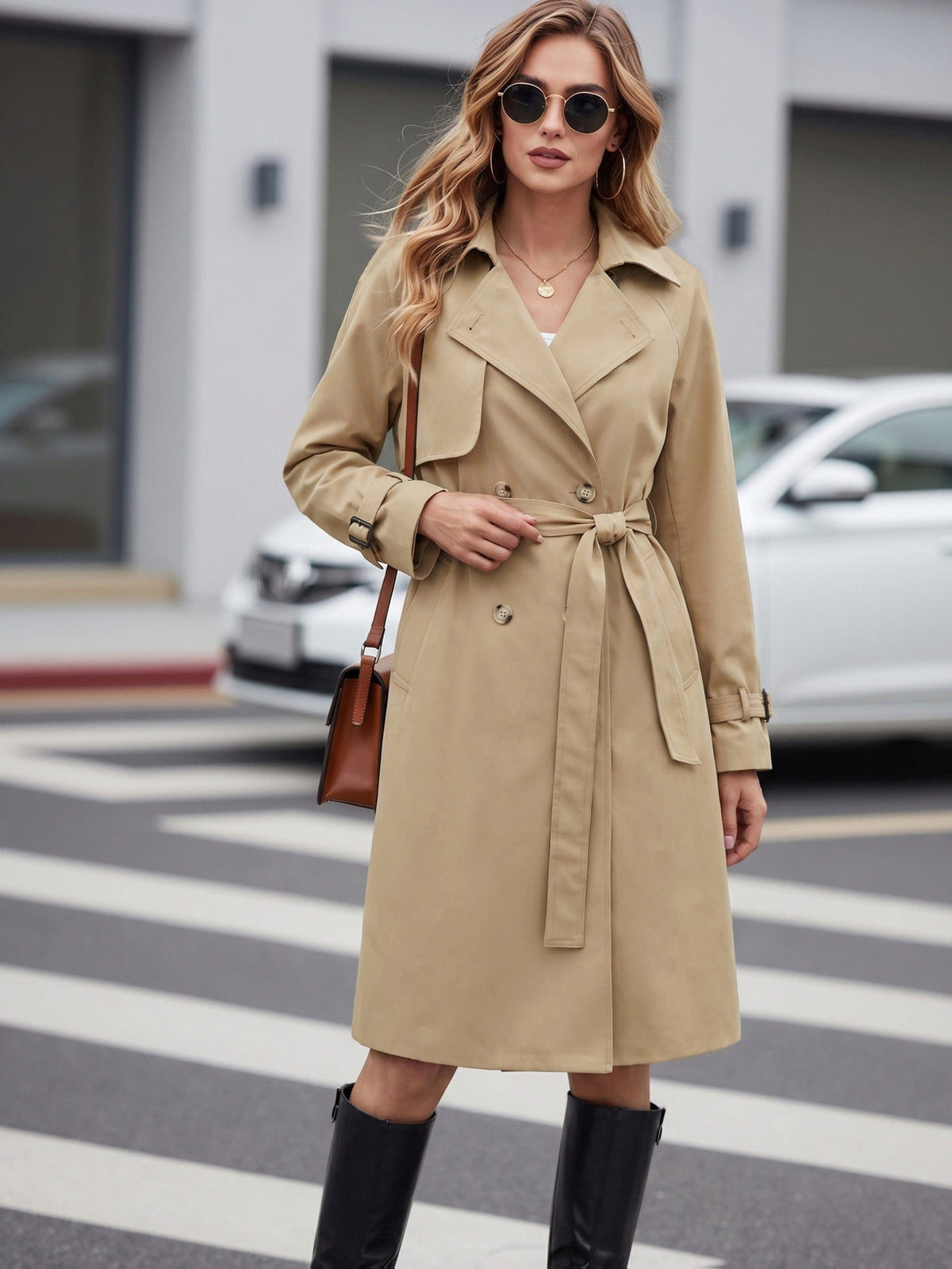 Woman wearing a belted trench coat with lapel collar and double breasted front, styled with black boots and a crossbody bag while crossing a city street._2