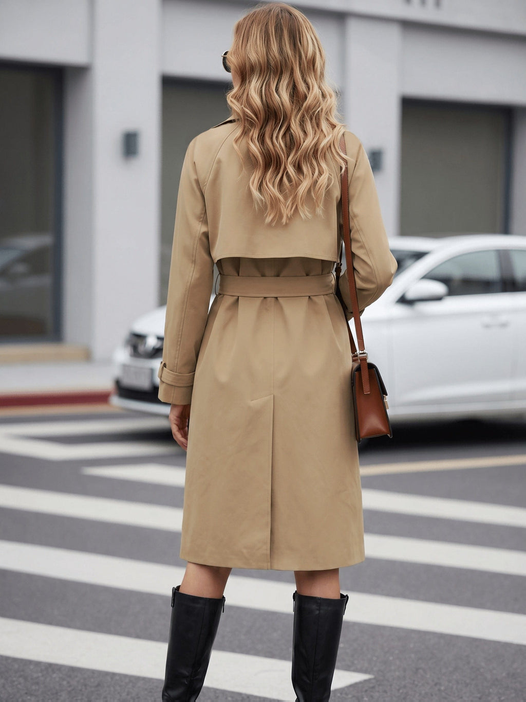 Woman wearing a belted trench coat with lapel collar and double breasted front, styled with black boots and a crossbody bag while crossing a city street._4