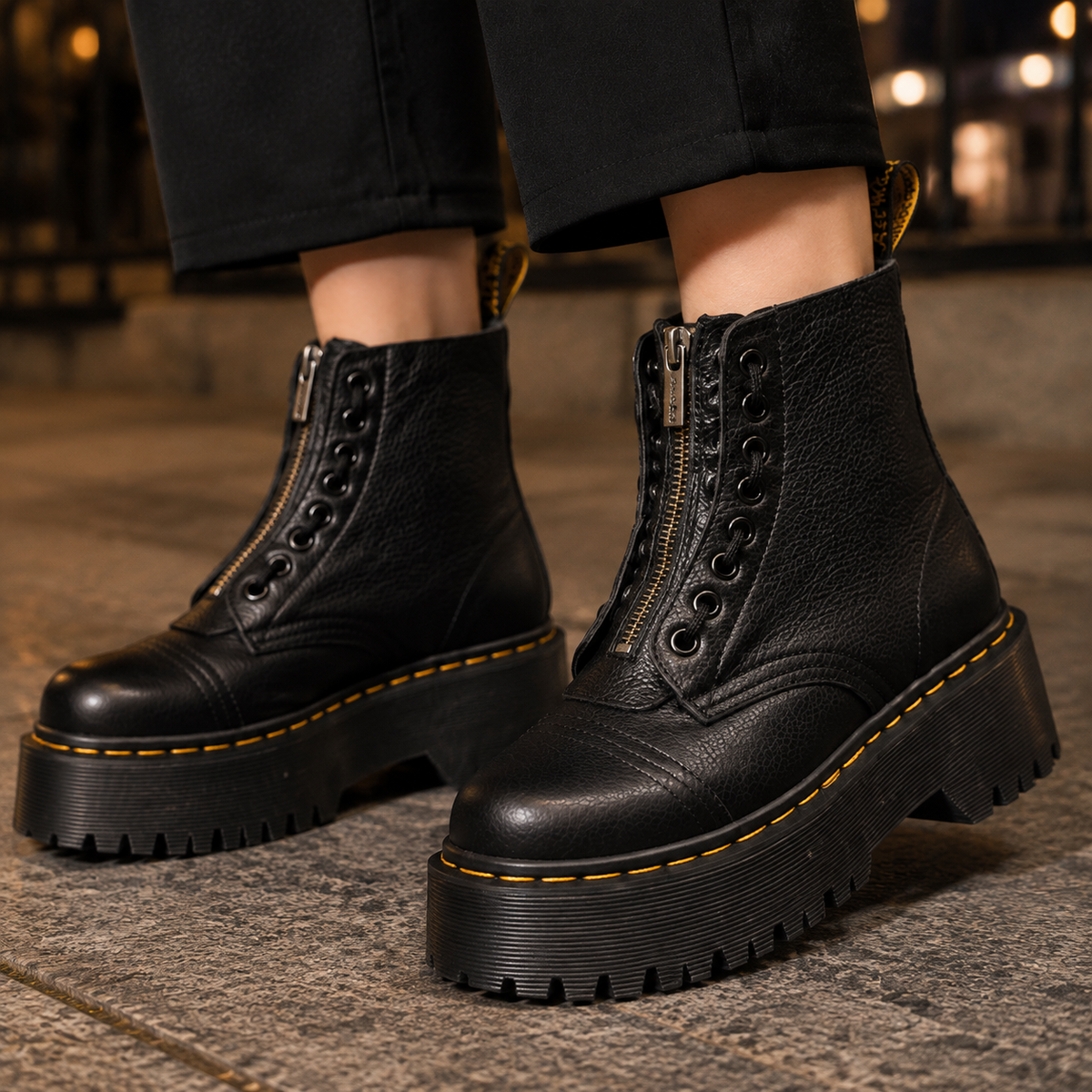 Pair of black ankle boots with a front zip, decorative eyelets, chunky platform lug sole and yellow contrast stitching worn with black cropped trousers._0
