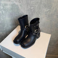 Pair of black ankle boots with buckle harness detail and low block heel displayed on a white pedestal against a textured grey backdrop._0