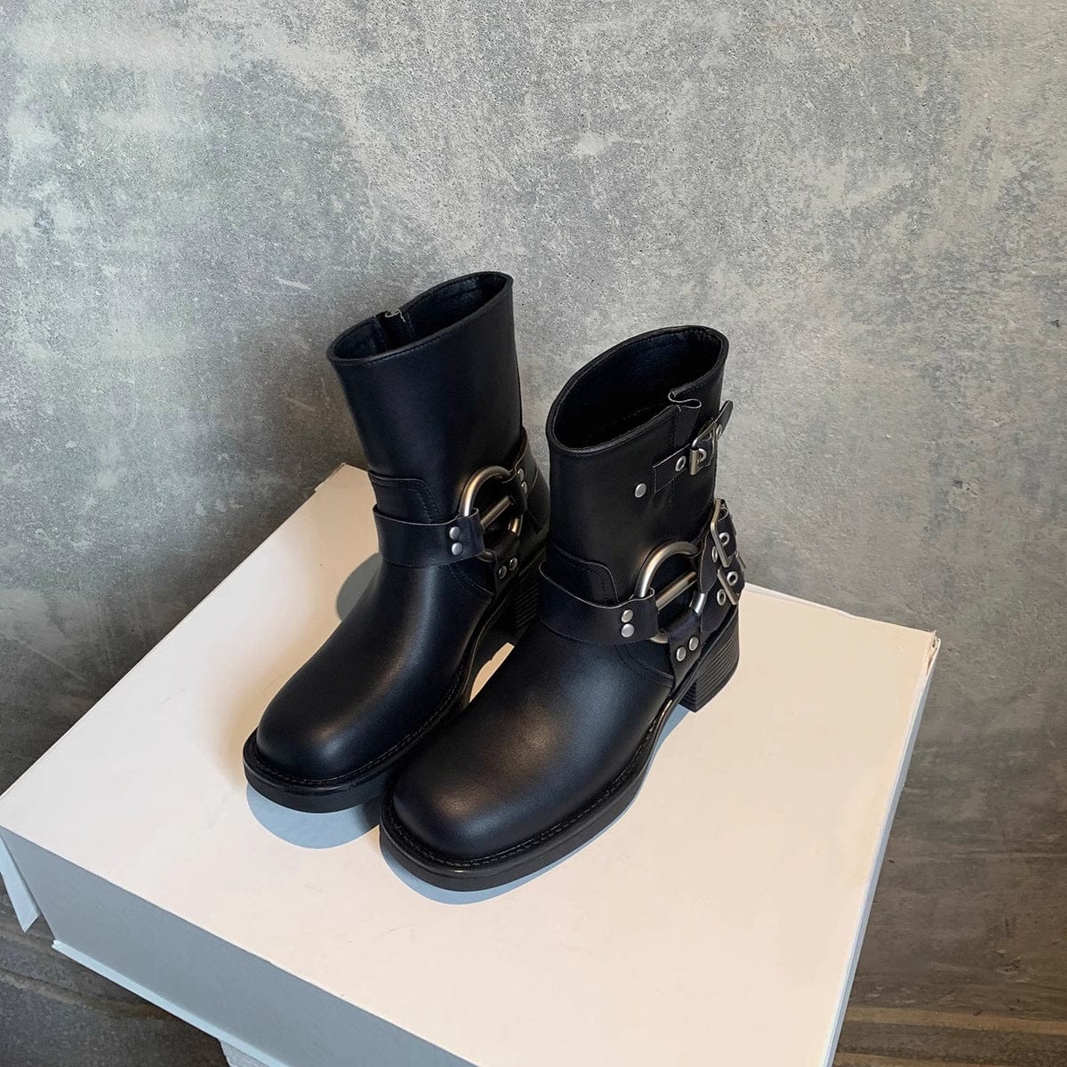 Pair of black ankle boots with buckle harness detail and low block heel displayed on a white pedestal against a textured grey backdrop._0