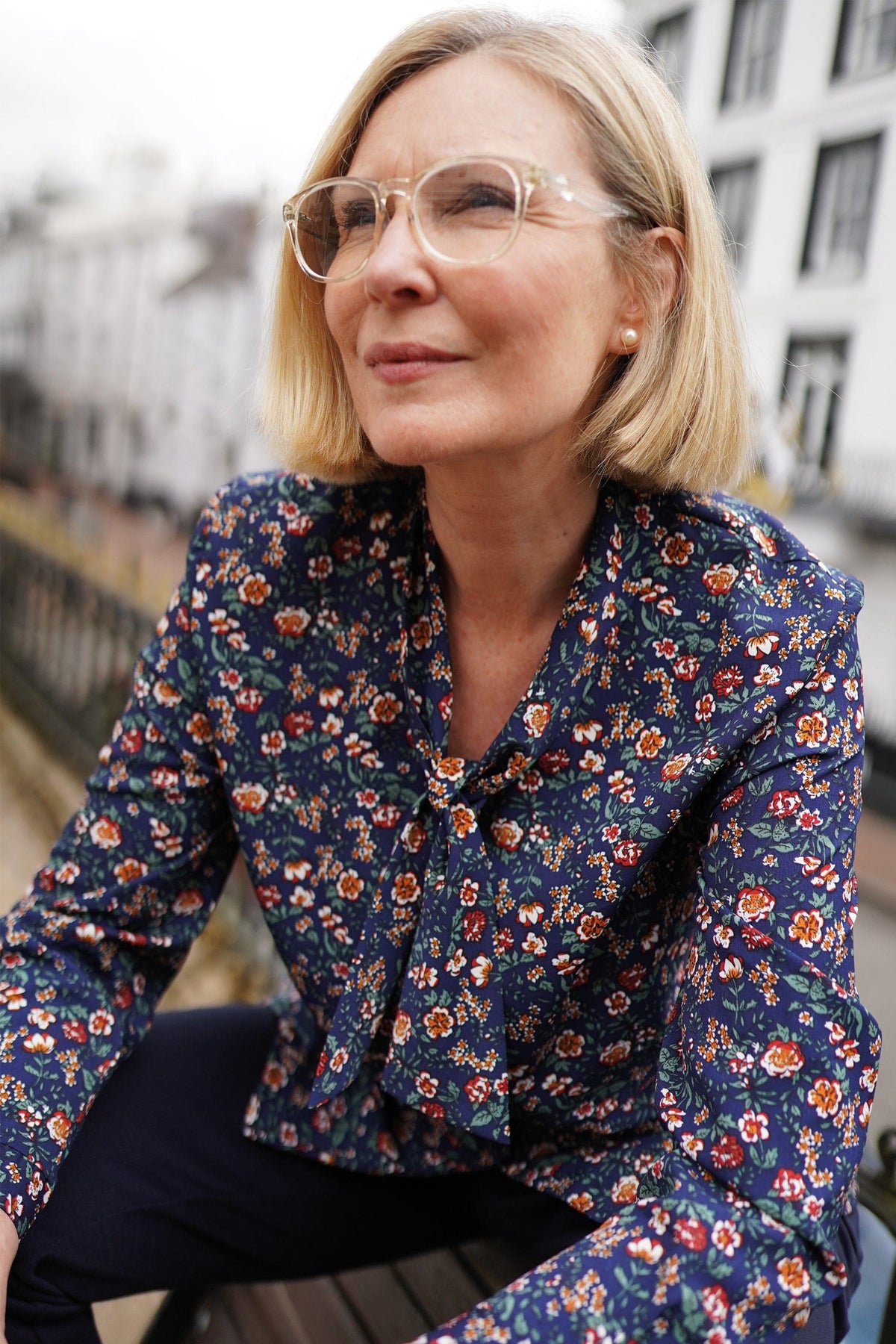 Woman in clear-framed glasses wearing a navy blouse with a small red, orange, cream and green floral print and a tied neckline, seated outdoors._0