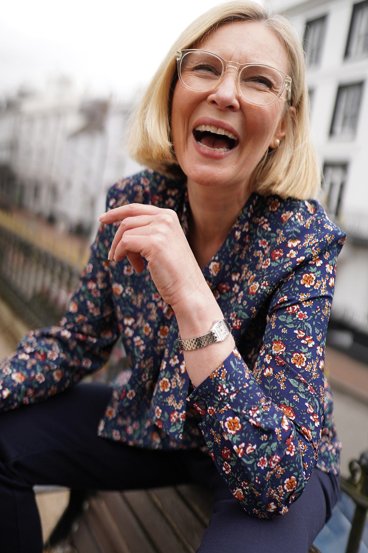 Woman in clear-framed glasses wearing a navy blouse with a small red, orange, cream and green floral print and a tied neckline, seated outdoors._1
