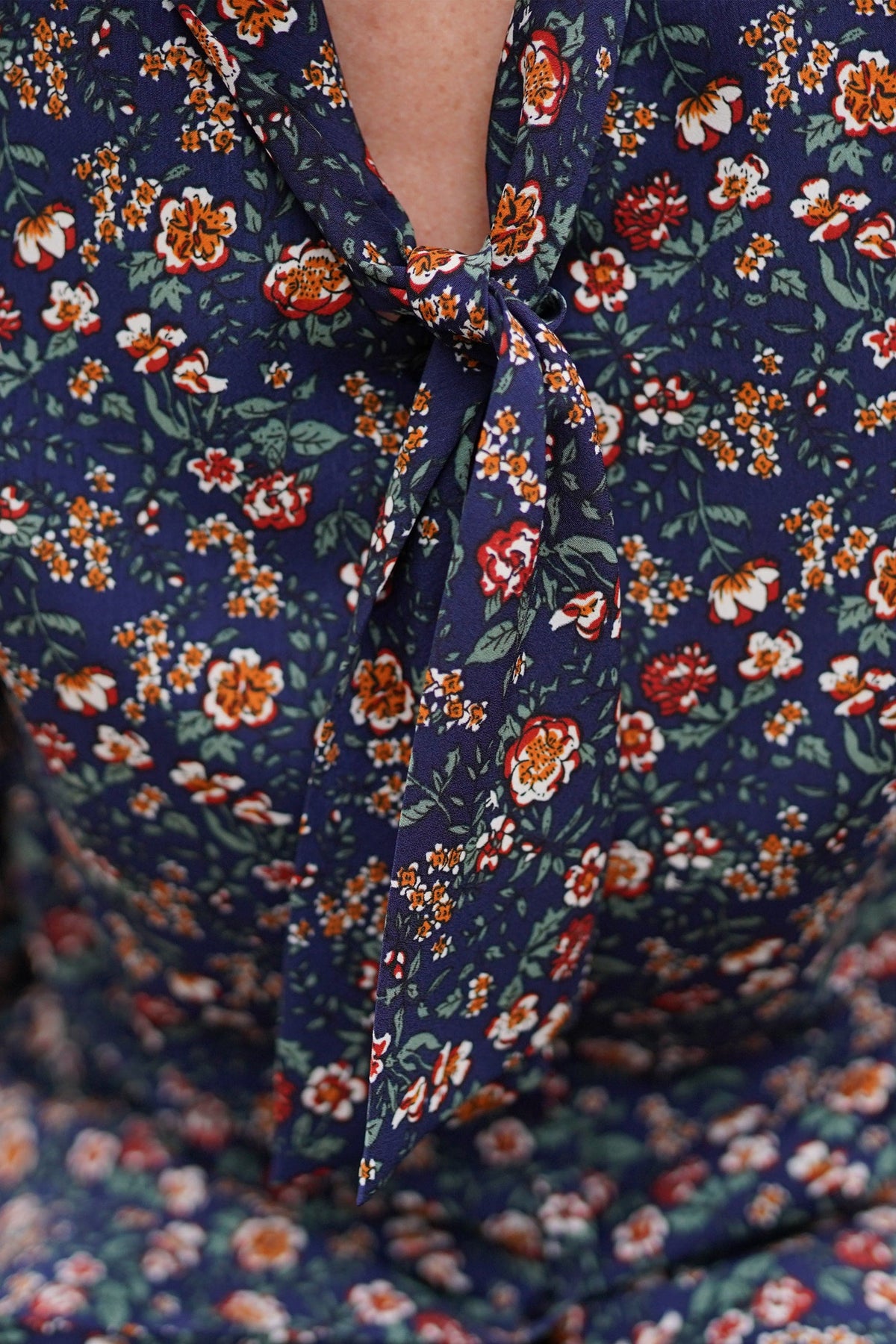 Woman in clear-framed glasses wearing a navy blouse with a small red, orange, cream and green floral print and a tied neckline, seated outdoors._2