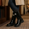 Person wearing glossy black knee-high rain boots with a folded black cuff, drawstring toggle and chunky lug sole, paired with dark jeans and a tan coat._0