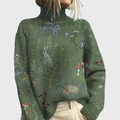 Woman wearing a loose green turtleneck sweater printed with wolves, a hare, birds, mushrooms and plants, paired with beige trousers._0