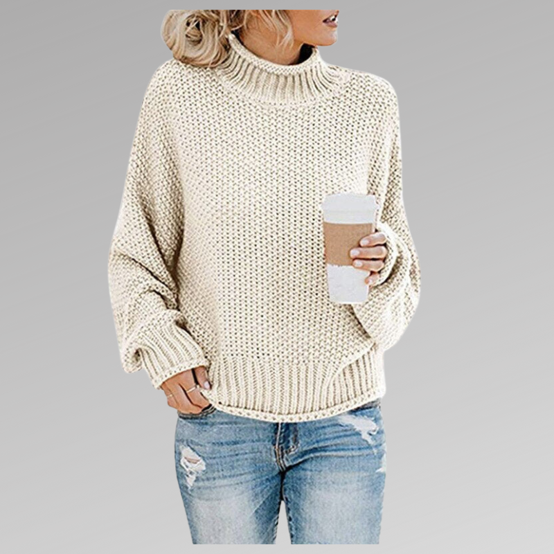 Woman wearing a chunky knit oversized turtleneck sweater in cream paired with distressed blue jeans while holding a coffee cup._0