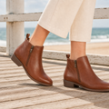 A person wearing tan ankle boots with side zips and low stacked heels, standing on a wooden boardwalk near the sea with cream wide-leg trousers._0