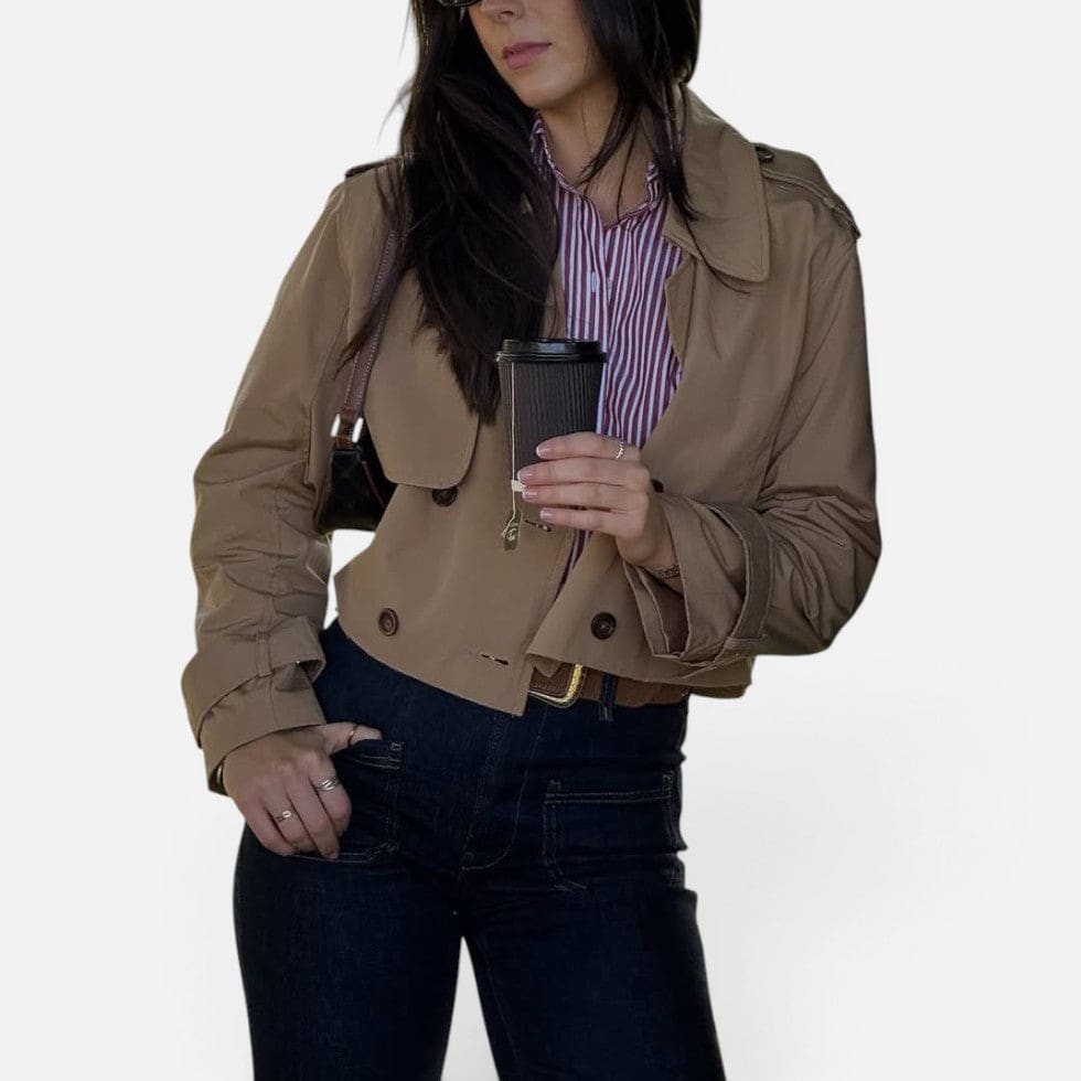 Cropped trench jacket in a neutral tone with double breasted button closure, notched lapel collar, shoulder epaulettes, and belted buckle cuffs displayed flat on a light background._9