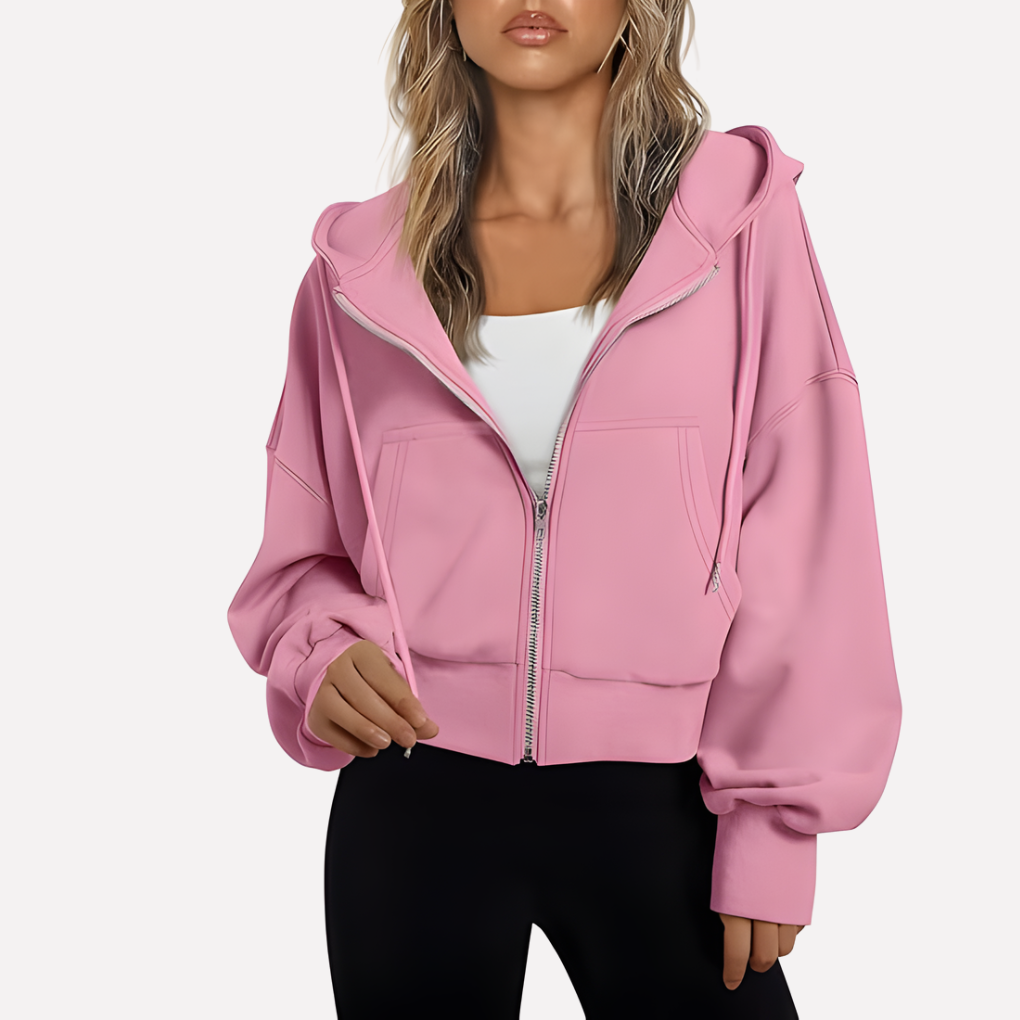 Blush pink cropped hoodie layered over a white top, with a drawstring hood, zip front, roomy sleeves, and front pockets.