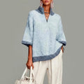 Woman wearing an oversized light blue knit sweater with darker blue trim, stand collar, short zip and three-quarter sleeves, with cream trousers and a woven bag._0
