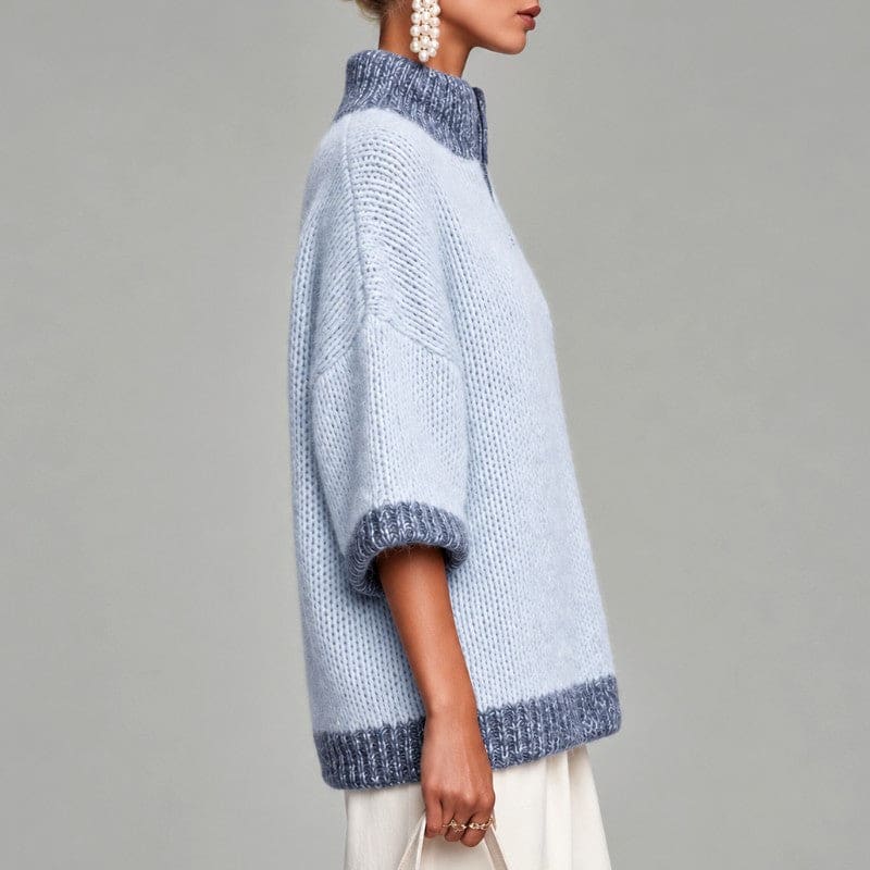 Woman wearing an oversized light blue knit sweater with darker blue trim, stand collar, short zip and three-quarter sleeves, with cream trousers and a woven bag._3