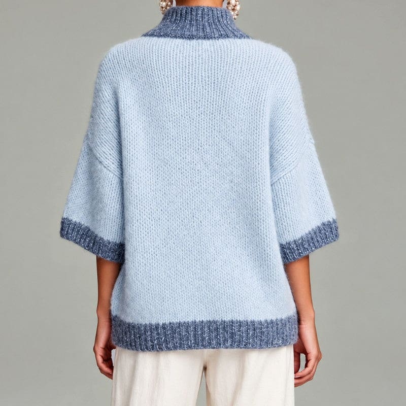 Woman wearing an oversized light blue knit sweater with darker blue trim, stand collar, short zip and three-quarter sleeves, with cream trousers and a woven bag._4