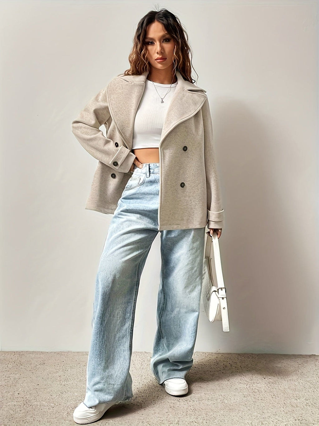 Woman wearing an oversized double-breasted pea coat with notched lapel, white crop top, and light wash jeans against a neutral background._3