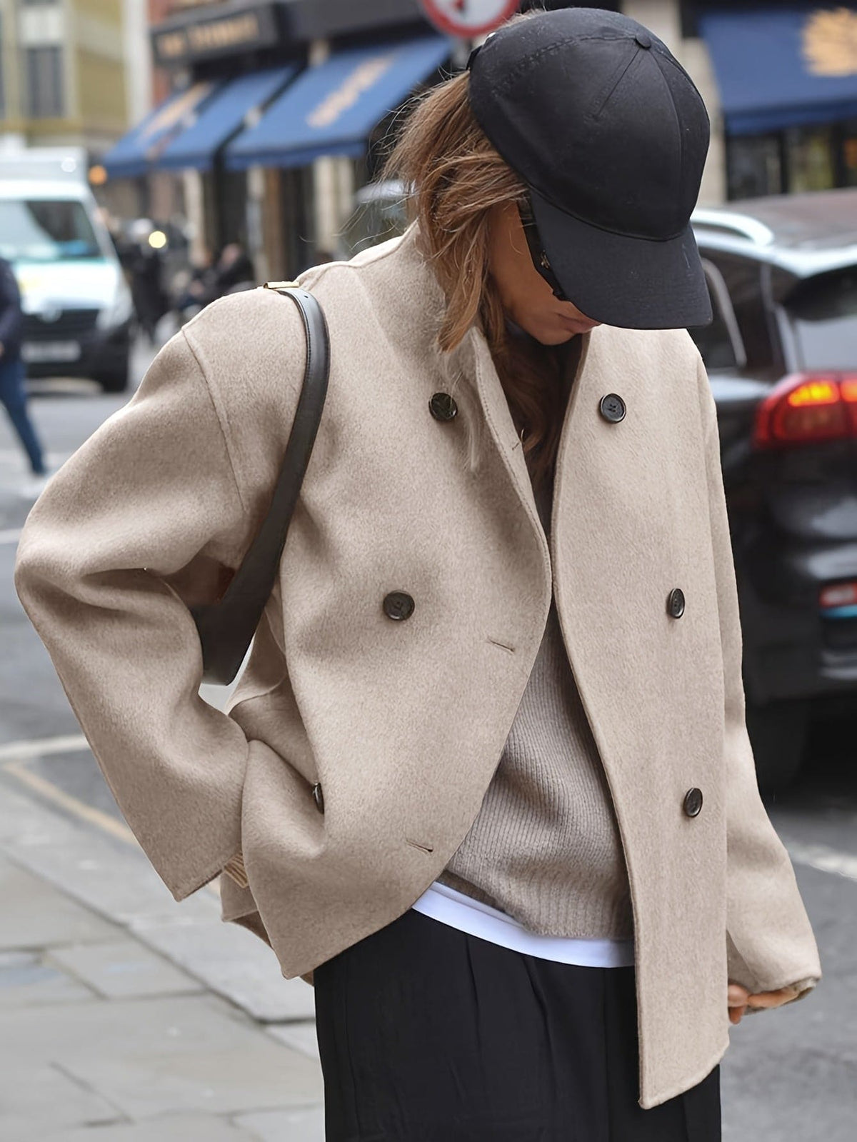 Woman wearing an oversized double breasted pea coat with stand collar and cropped silhouette, styled with a baseball cap on a city street._0