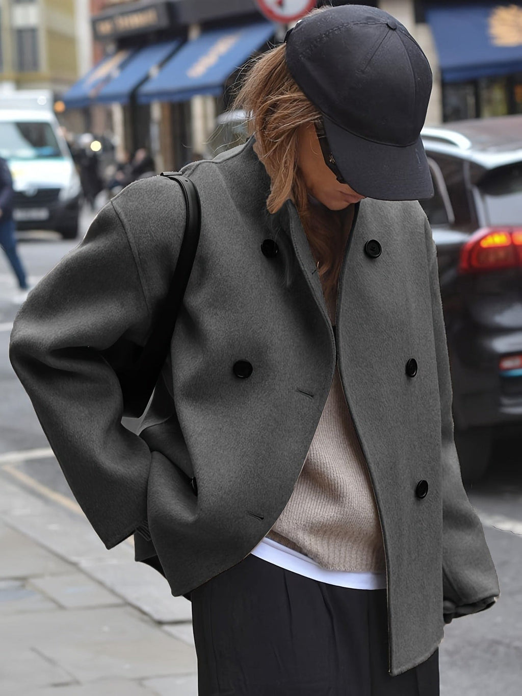 Woman wearing an oversized double breasted pea coat with stand collar and cropped silhouette, styled with a baseball cap on a city street._1