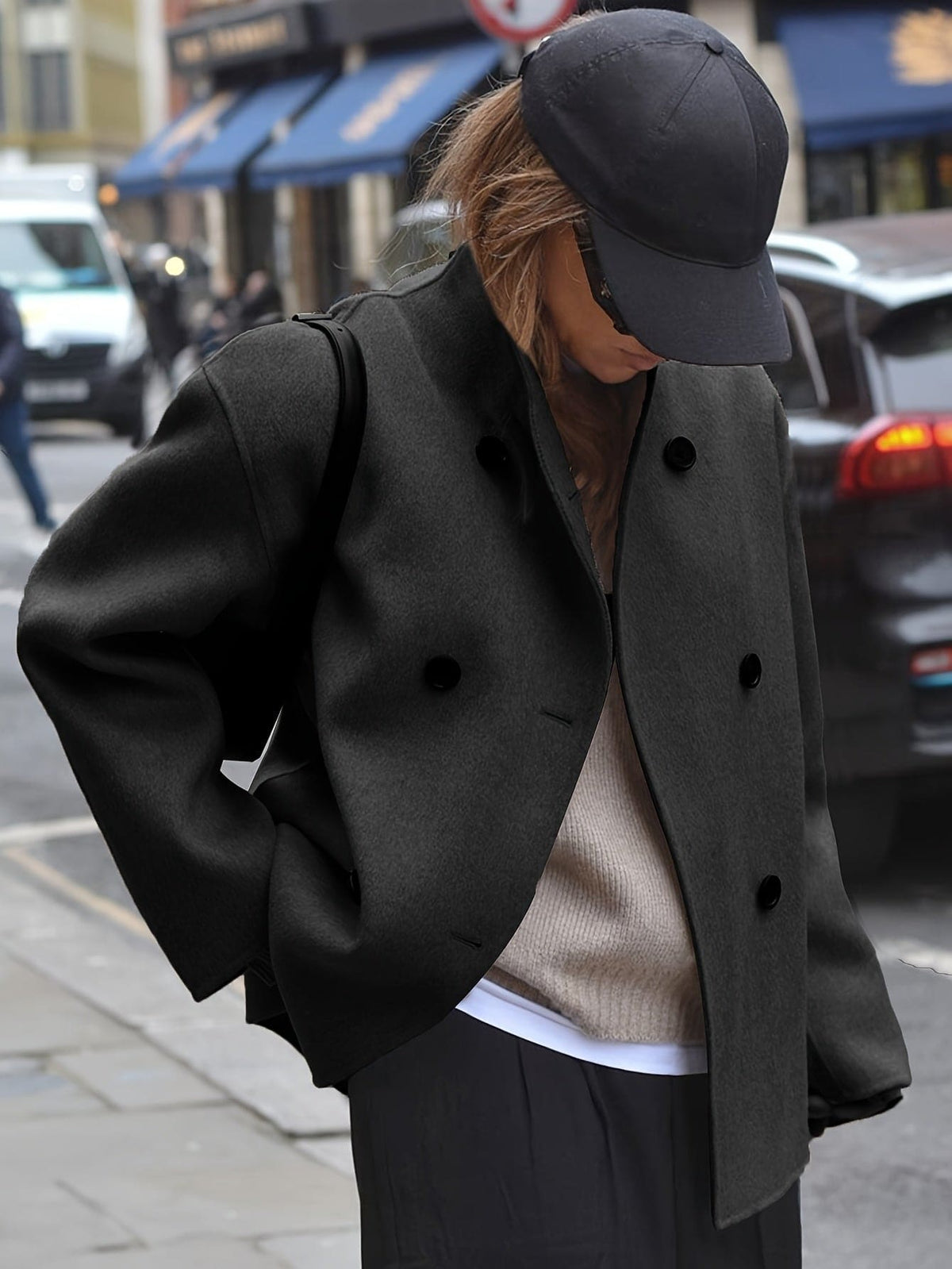 Woman wearing an oversized double breasted pea coat with stand collar and cropped silhouette, styled with a baseball cap on a city street._4