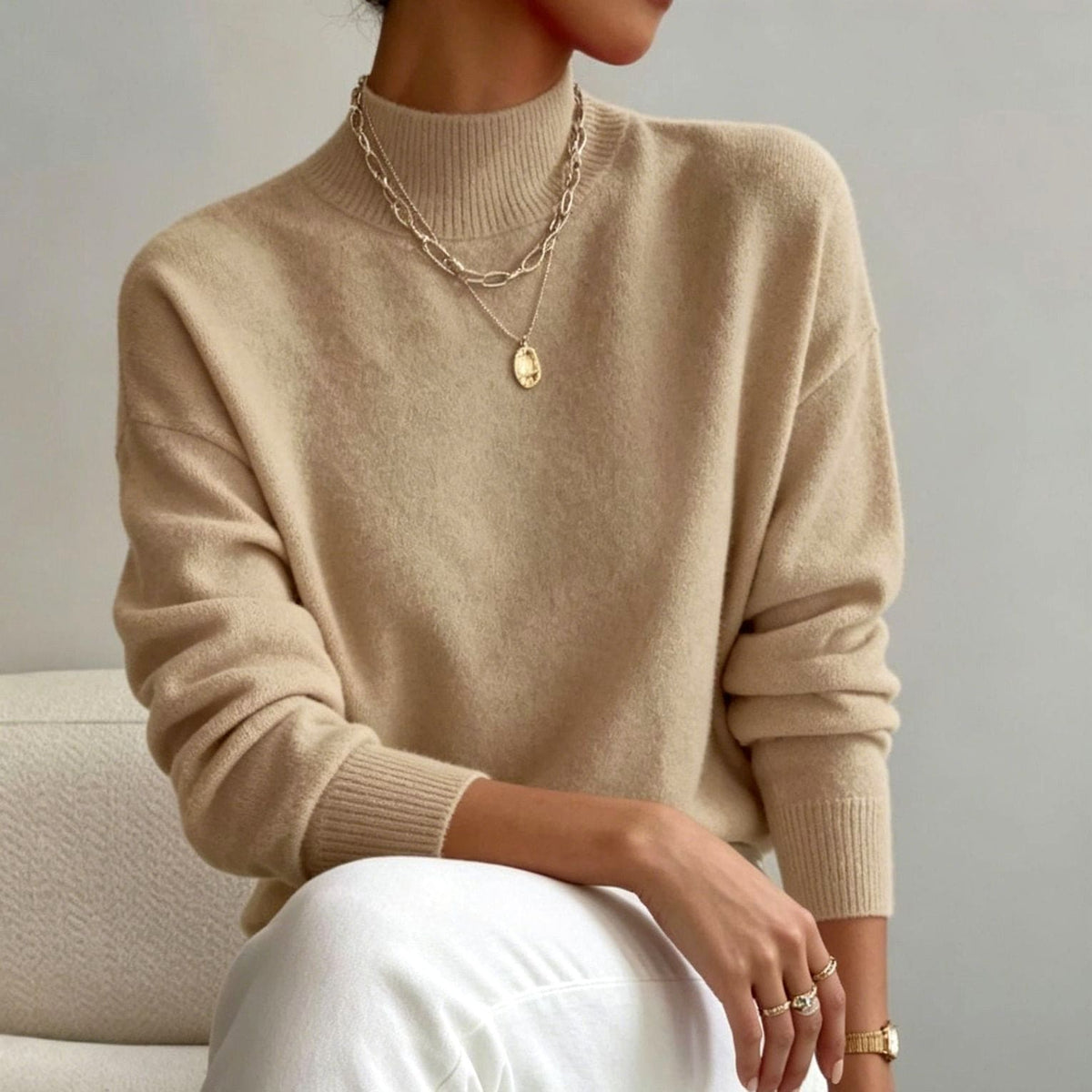 Woman seated wearing a beige mock neck sweater with dropped shoulders and ribbed cuffs, white trousers and layered gold necklaces._0