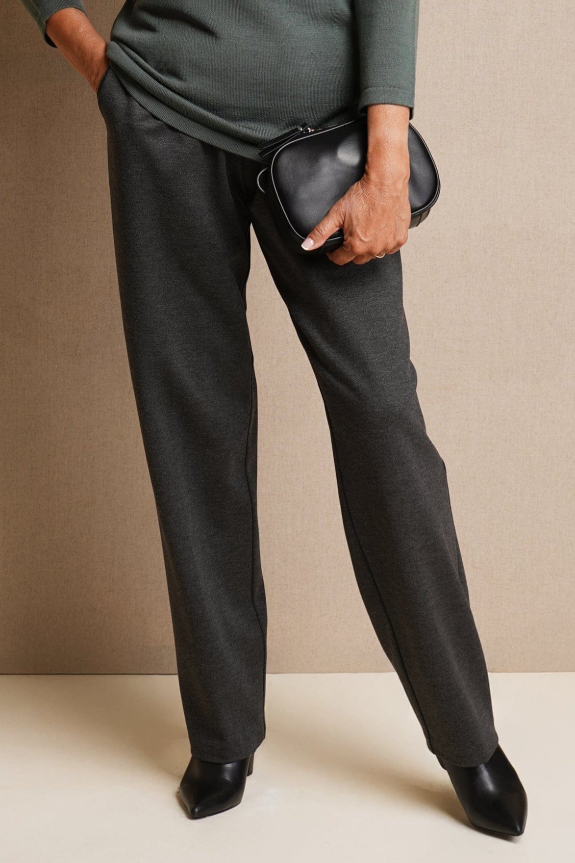 Woman wearing charcoal grey straight-leg trousers with a green knit top, black ankle boots and a small black bag._0