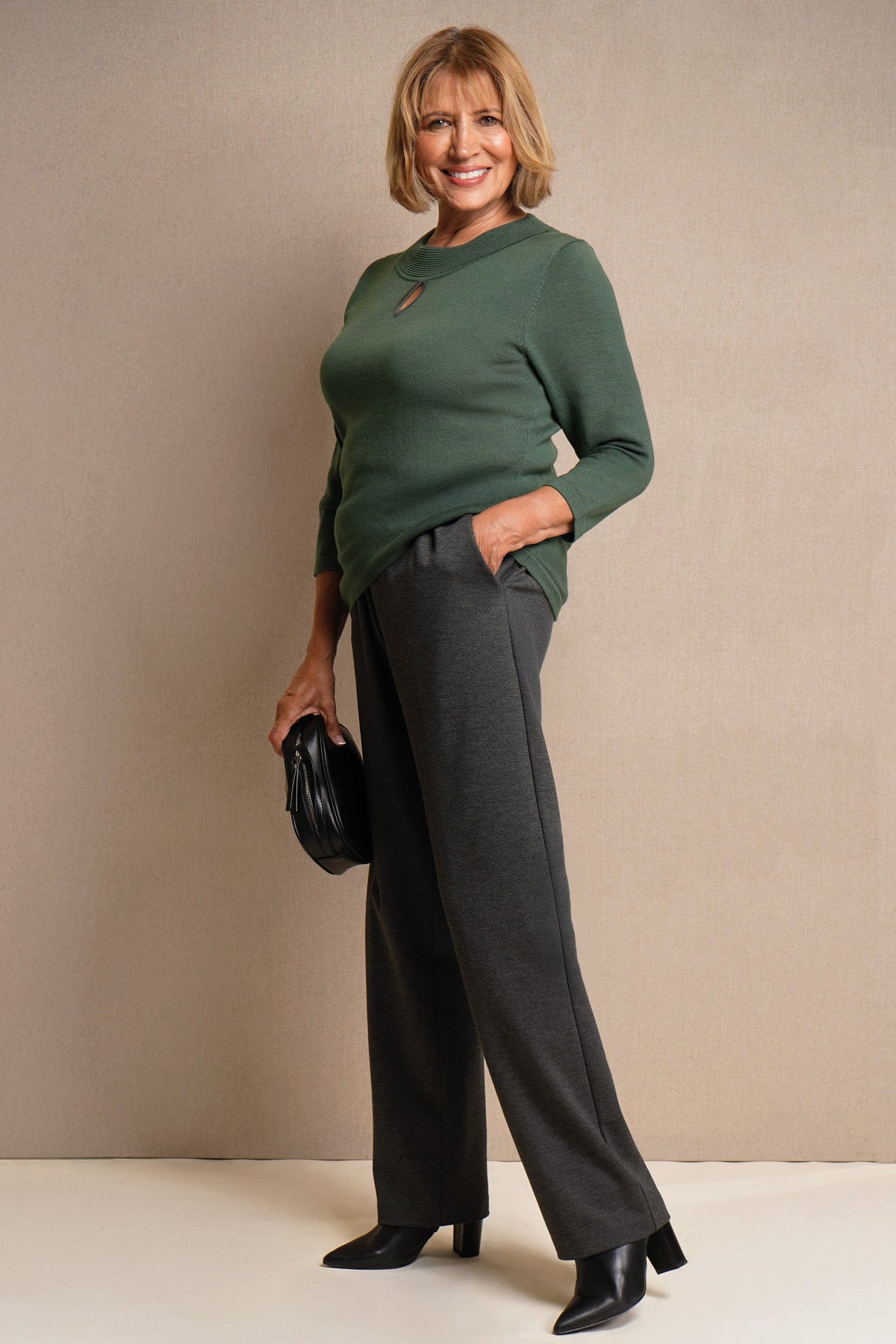Woman wearing charcoal grey straight-leg trousers with a green knit top, black ankle boots and a small black bag._1