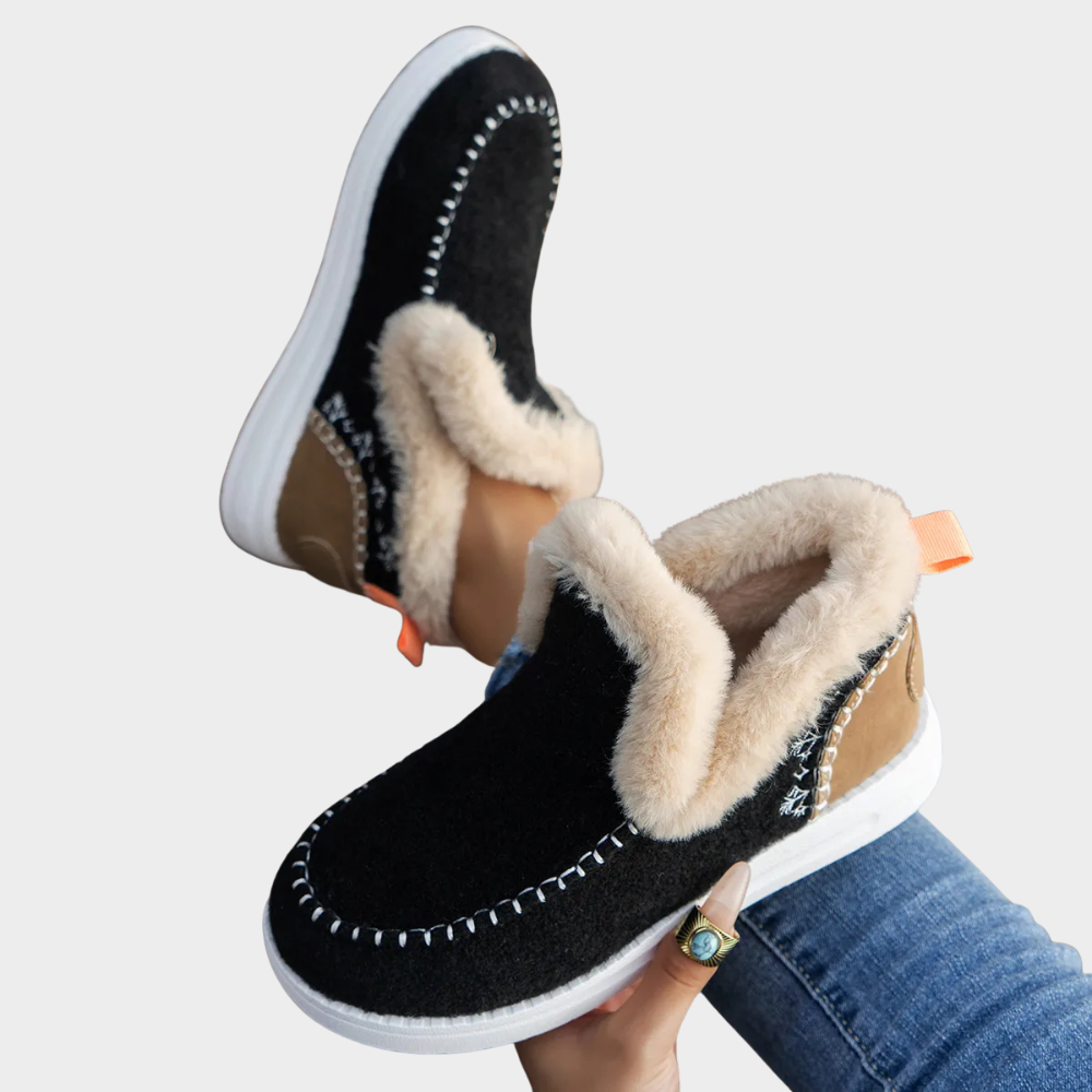 Woman holding a black ankle boot with faux fur lining and contrast tan heel panel, featuring white contrast stitching and a white sole._0