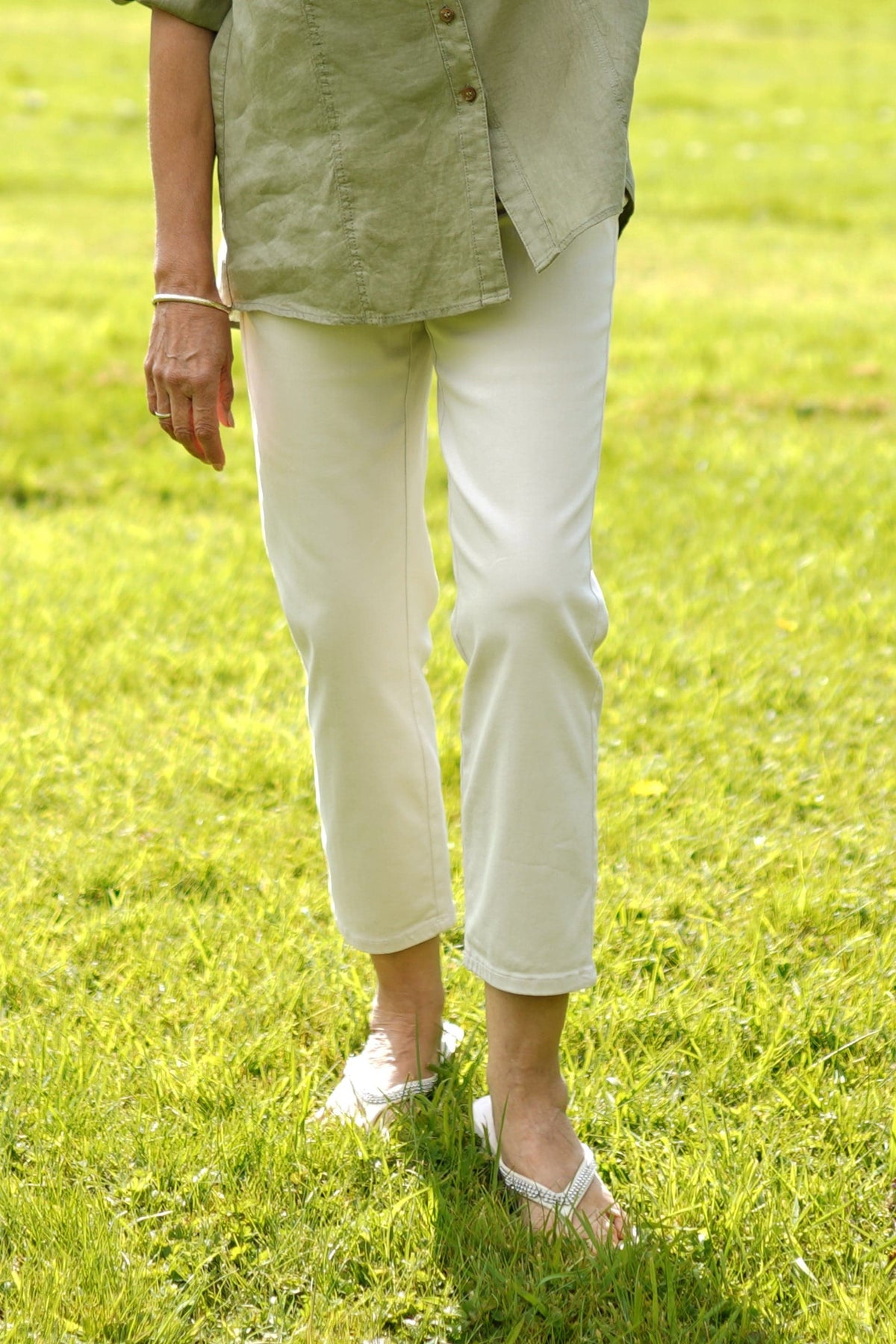 Woman standing on grass wearing cropped beige straight-leg trousers with a green short-sleeve button shirt and white sandals._0