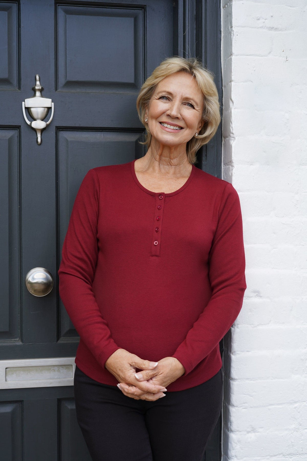 Smiling woman with short blonde hair wearing a burgundy long sleeve top with a round neck and five button front placket, standing in front of a dark door._2