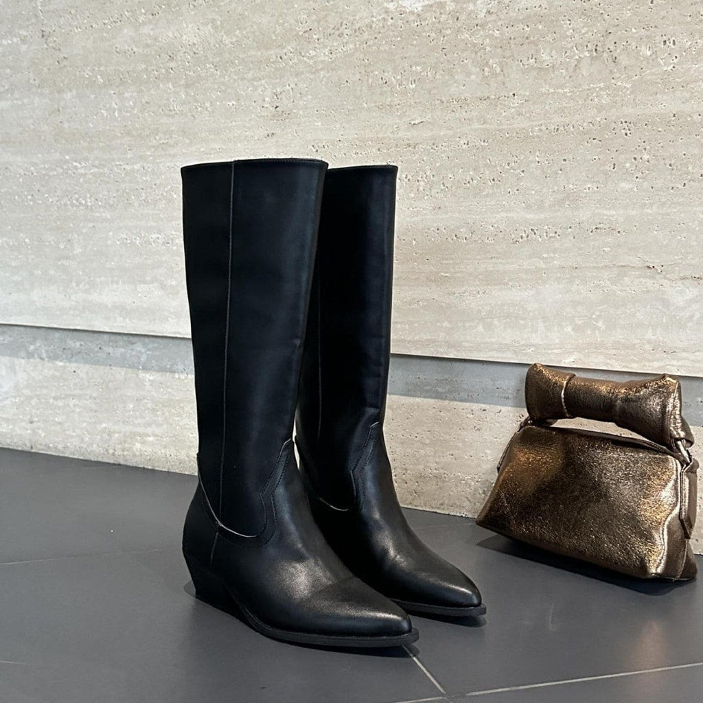 High-rise boots with pointed toe and sleek silhouette displayed on a dark surface with a handbag in the background._0