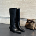 High-rise boots with pointed toe and sleek silhouette displayed on a dark surface with a handbag in the background._0