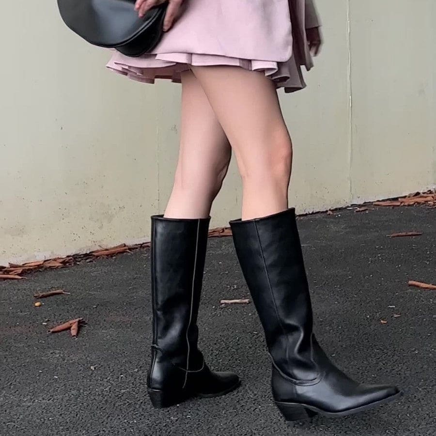High-rise boots with pointed toe and sleek silhouette displayed on a dark surface with a handbag in the background._2