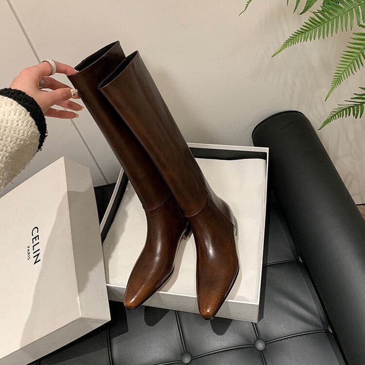 High-rise boots with pointed toe and sleek silhouette displayed on a dark surface with a handbag in the background._4