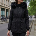Woman wearing a hooded parka jacket with faux fur trim, cinched waist, and flap pockets, standing outdoors on a city street._0