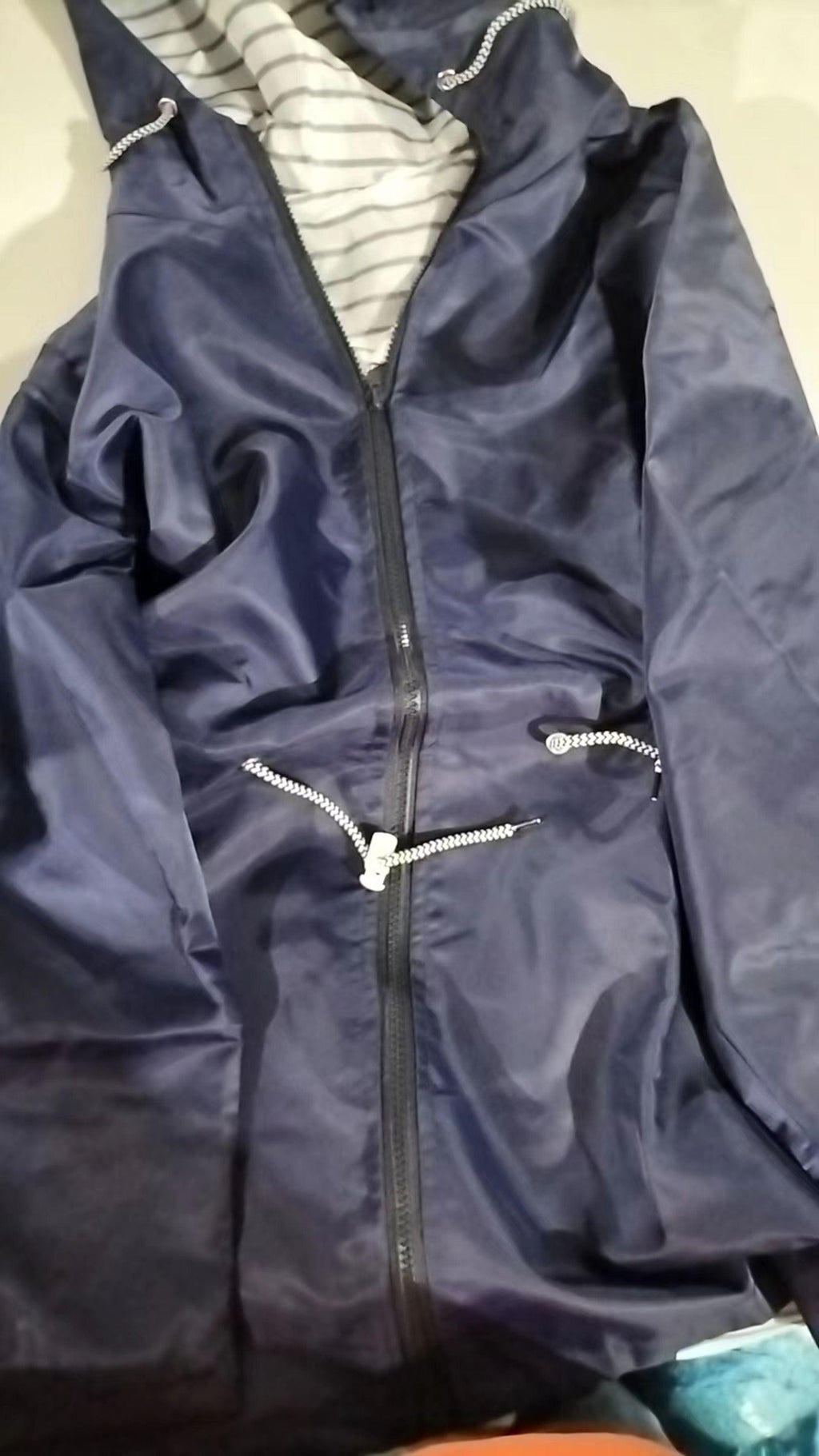 Close-up of a navy hooded rain coat lying flat, featuring the zipped front, raised hood, striped lining, and zippered pockets.