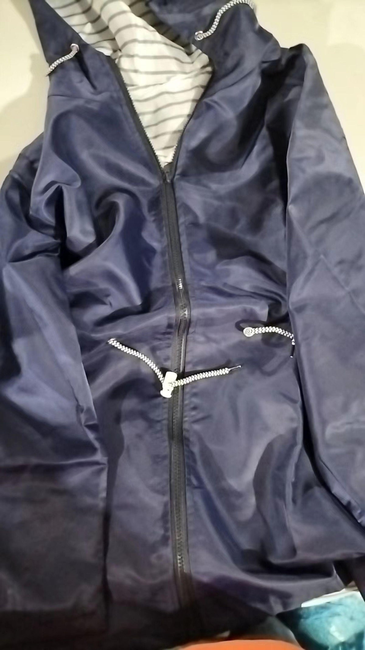 Close-up of a navy hooded rain coat lying flat, featuring the zipped front, raised hood, striped lining, and zippered pockets.
